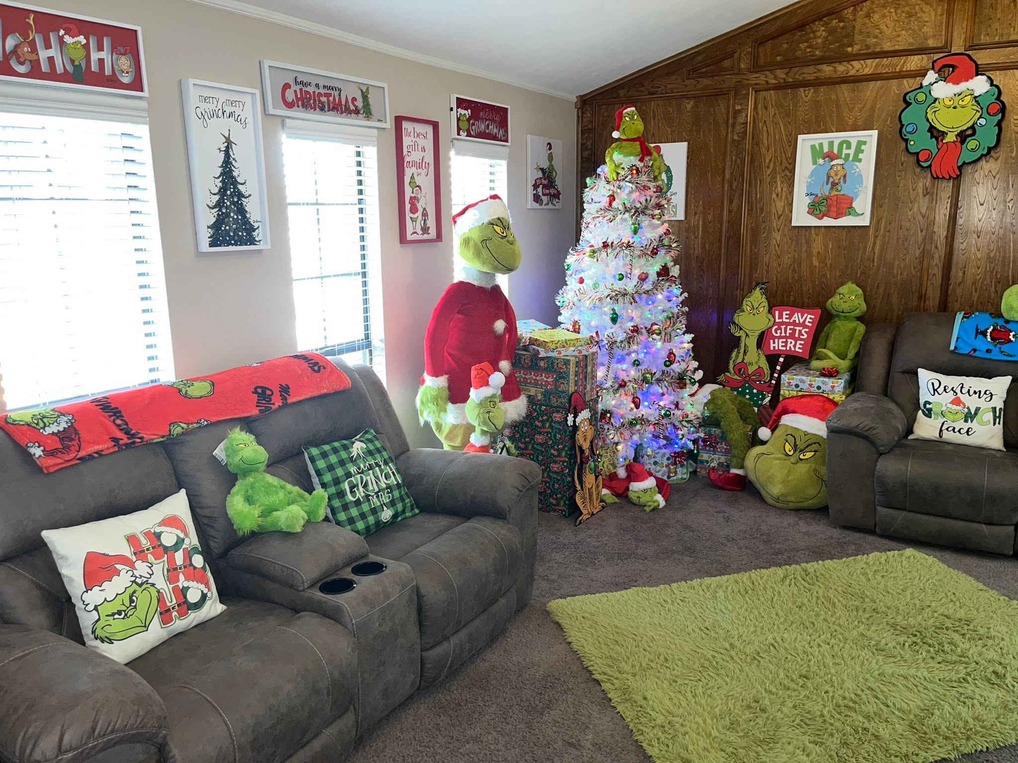 Living room decorated for Christmas with Grinch-themed pillows, plushies, wall art, and a snowy tree.