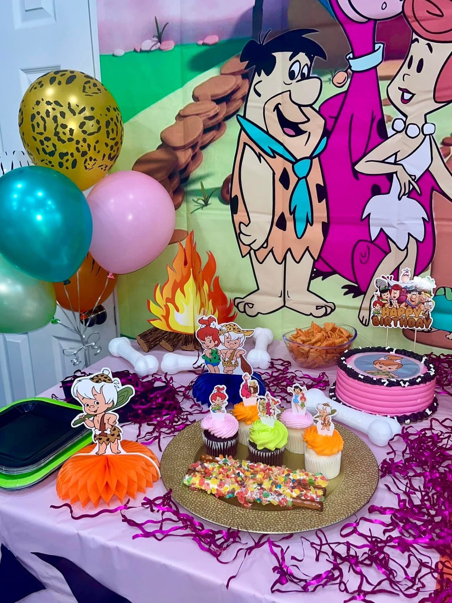 Flintstones themed birthday party table with cupcakes, festive decorations, and character cutouts.