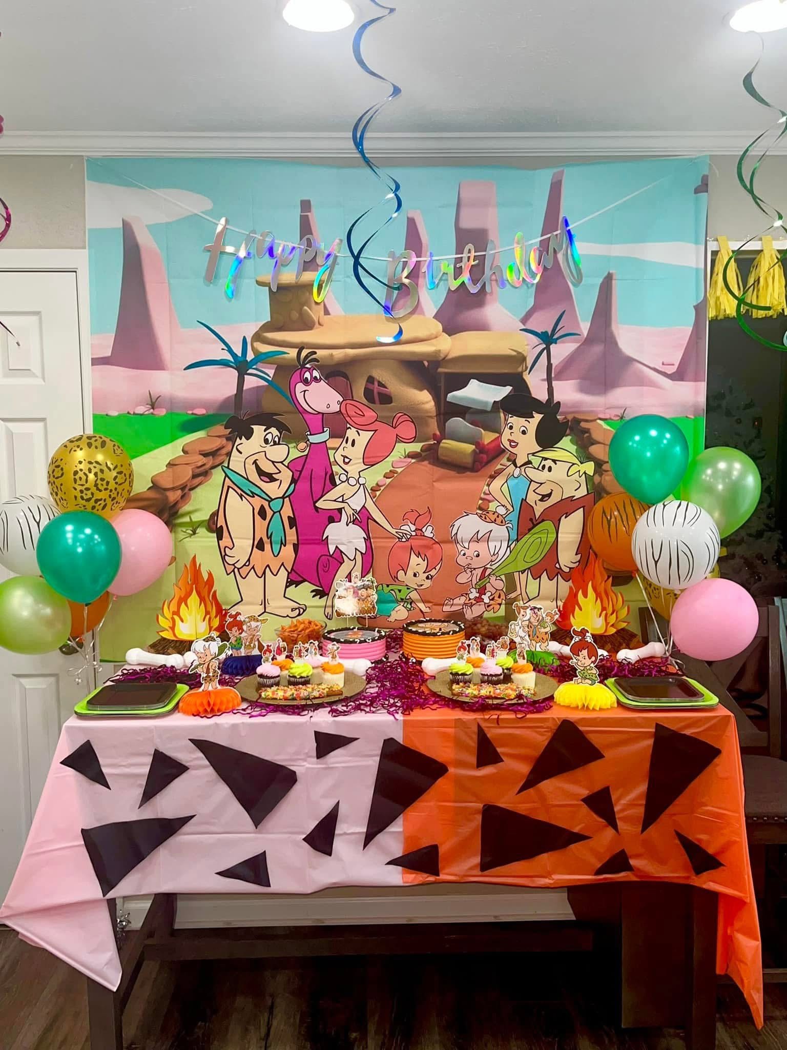 The Flintstones themed birthday party table setup with a decorative backdrop, green and pink balloons, and cartoon-themed snacks.