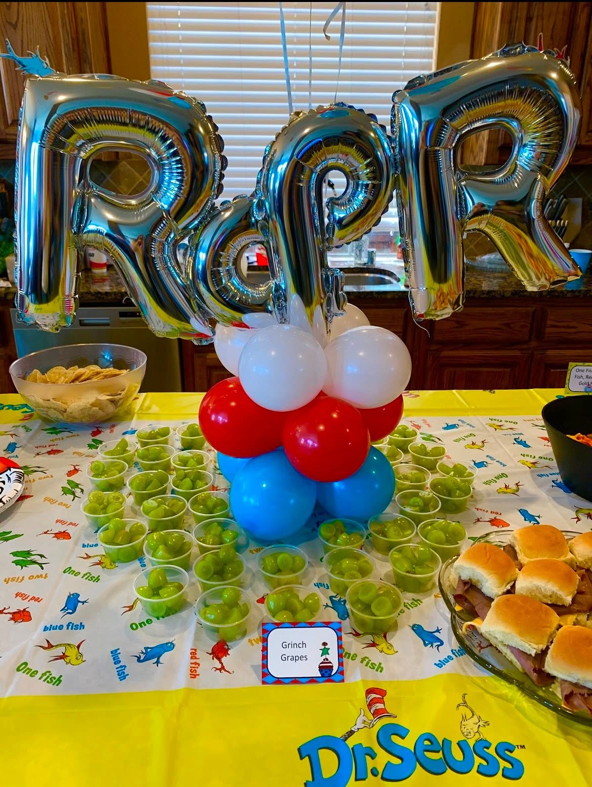 Dr. Seuss themed party table with Grinch Grapes cups, slider sandwiches, and silver ROR balloon decor.