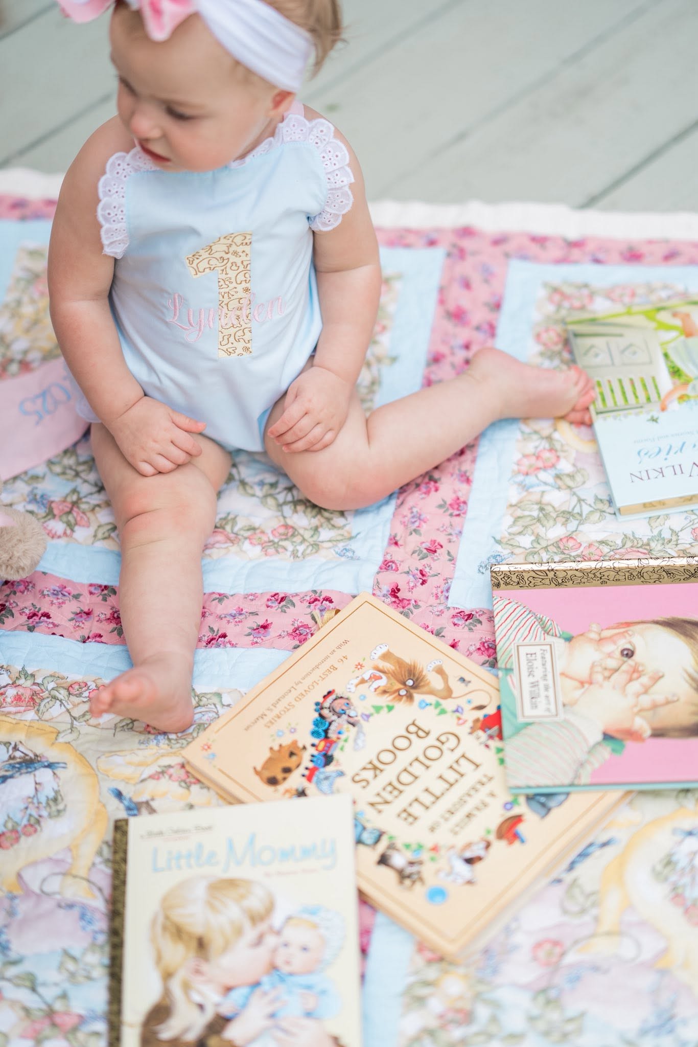 Baby girl wearing a first birthday onesie sitting on a patchwork quilt surrounded by classic storybooks.