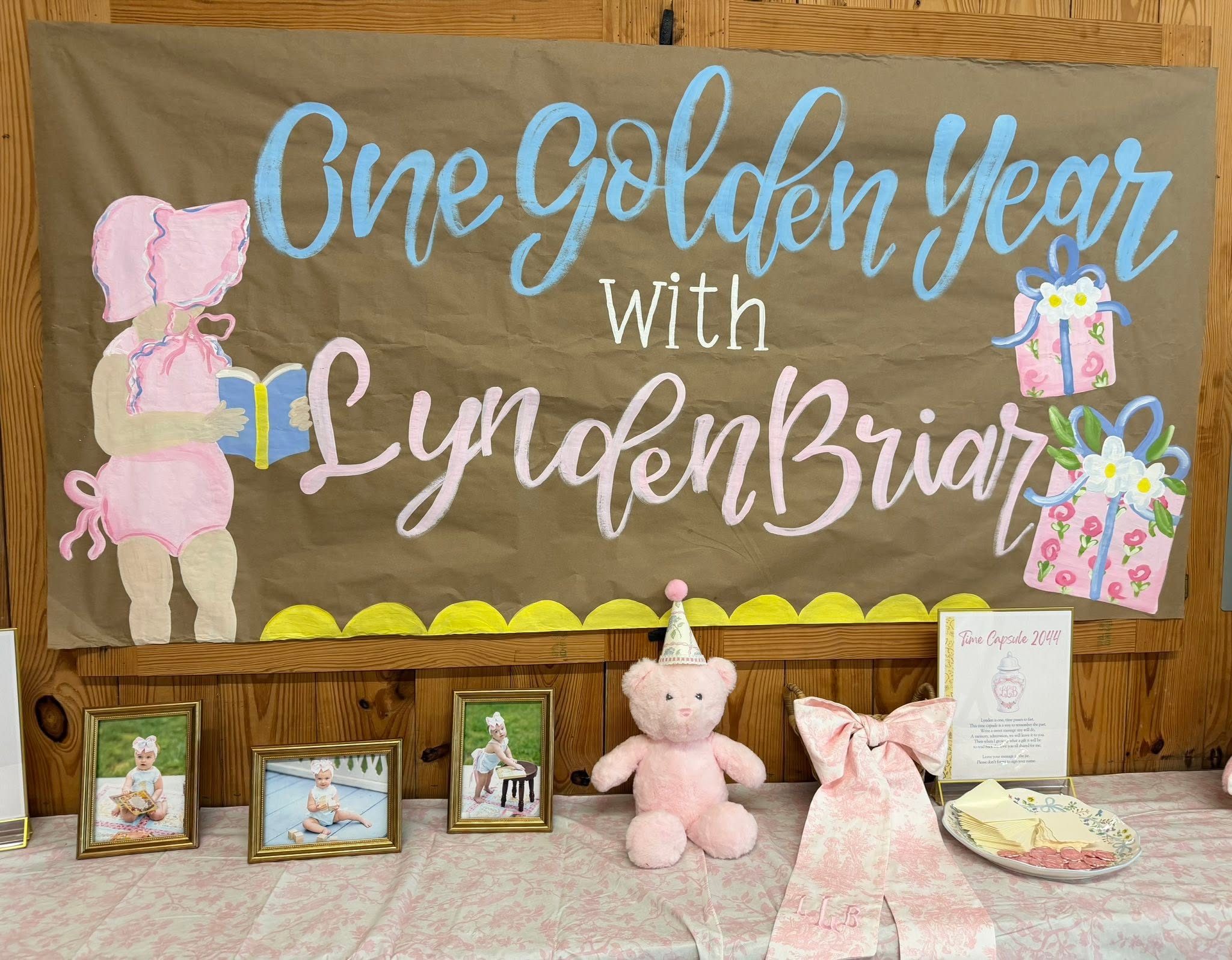 First birthday party decoration featuring a hand-painted banner saying One Golden Year with LyndenBriar, photos, and a pink teddy bear.
