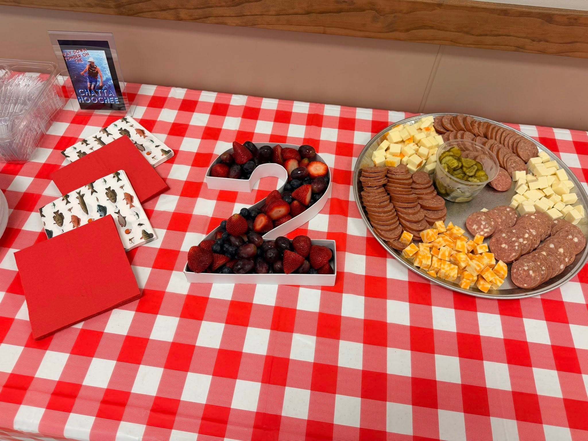 Table with red and white checkered tablecloth featuring a number 2 snack bin filled with berries and a charcuterie tray.