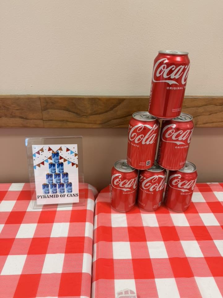 A pyramid of six Coca-Cola cans stacked on a red and white checkered tablecloth next to a small instructional sign.