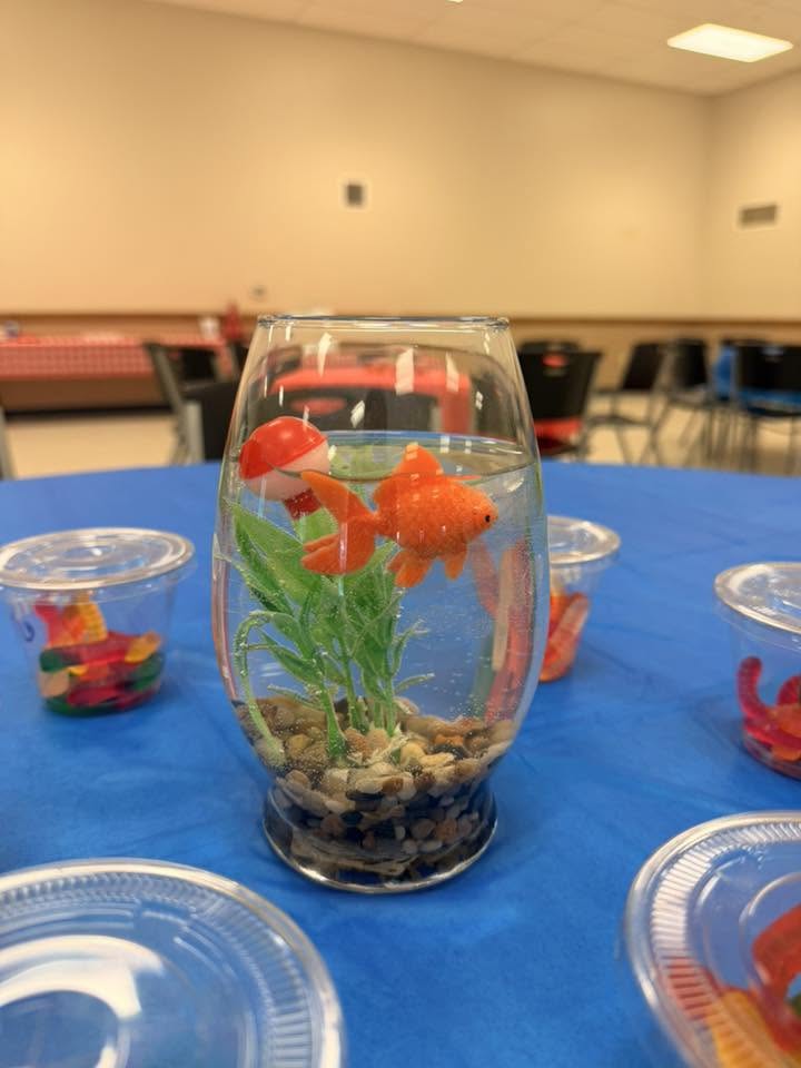 Glass vase table centerpiece with a toy goldfish, green plant, red bobber, and pebbles on a blue tablecloth.