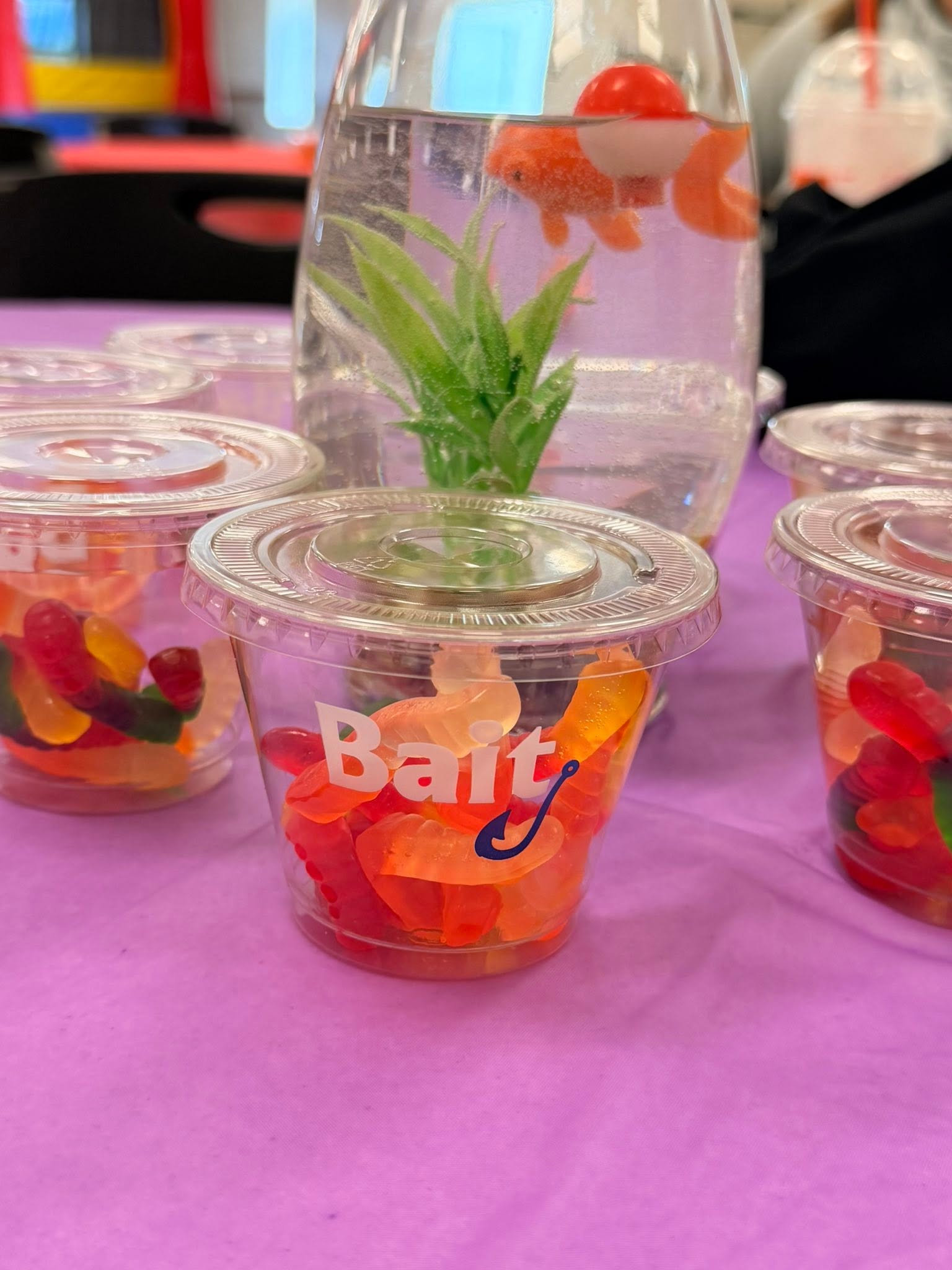 Clear plastic party cups labeled Bait filled with gummy worms on a purple tablecloth