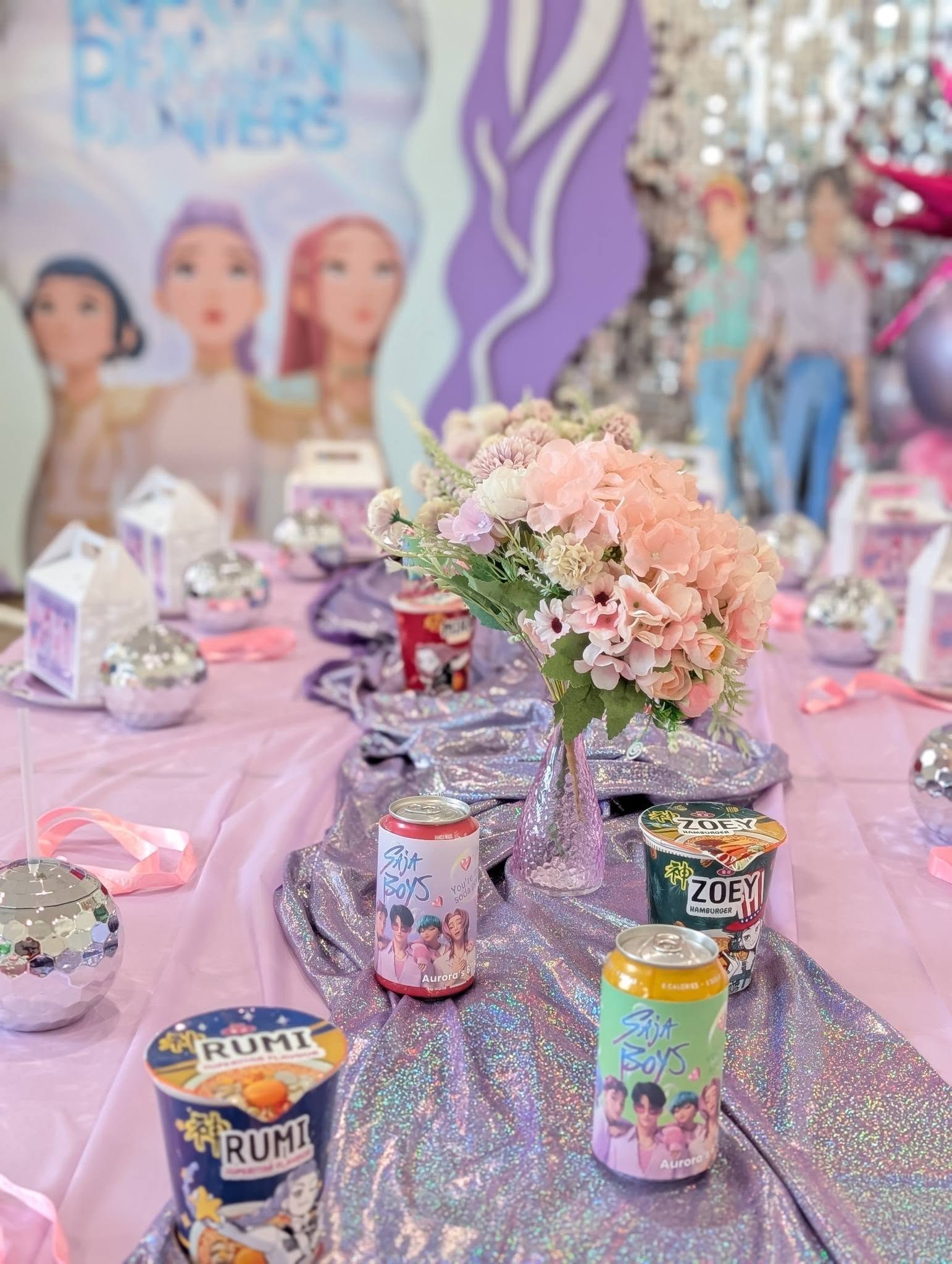 Table decorated with purple glitter runner, floral centerpiece, and Saja Boys themed soda cans and ramen cups.