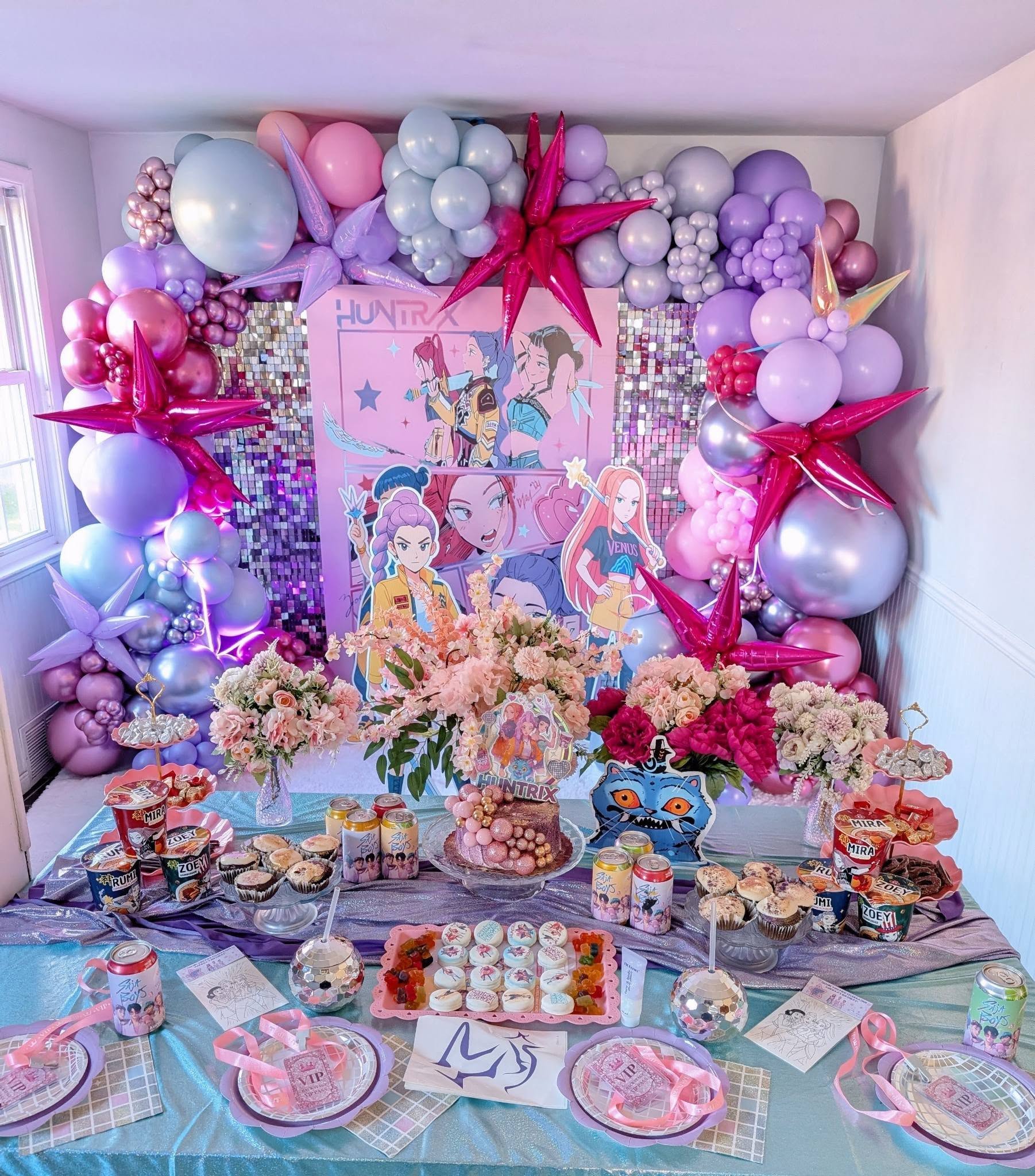 Table at a party themed after anime characters featuring custom drink cans, treats, and a balloon garland backdrop.