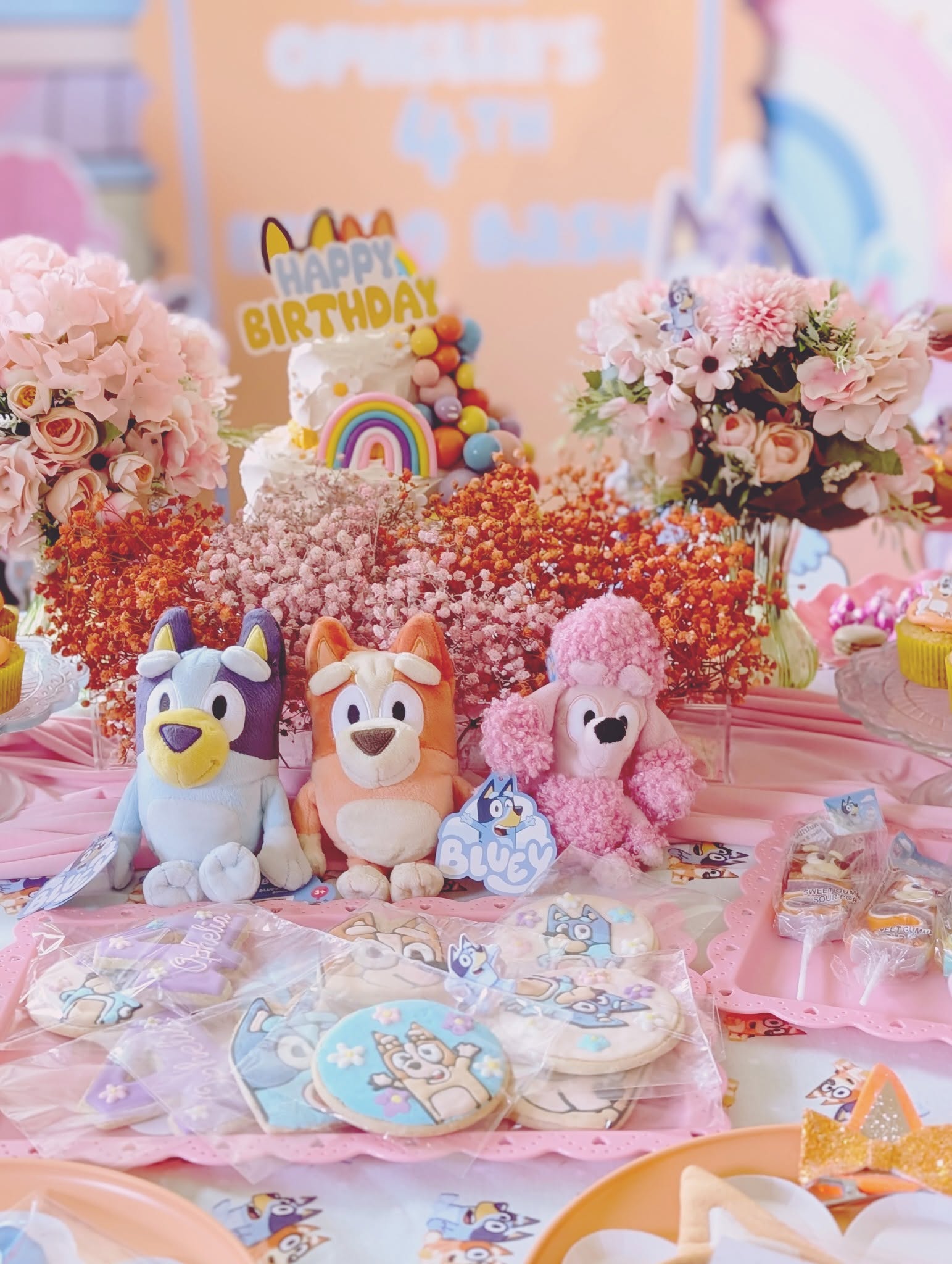 Bluey and Bingo plush toys with floral centerpieces and decorated birthday cookies on a party table.
