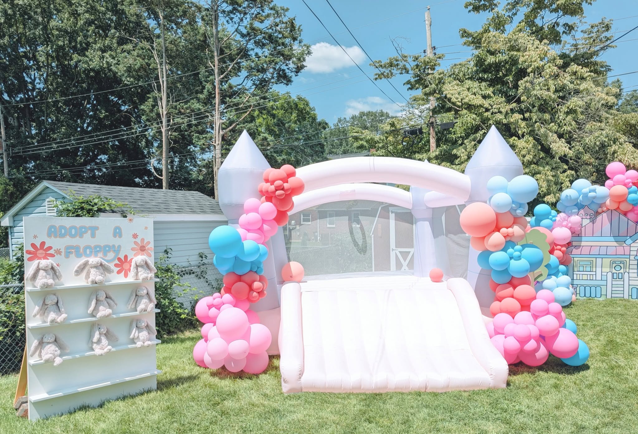White inflatable bounce house with slide and pastel balloon arch next to a plush bunny adoption display.