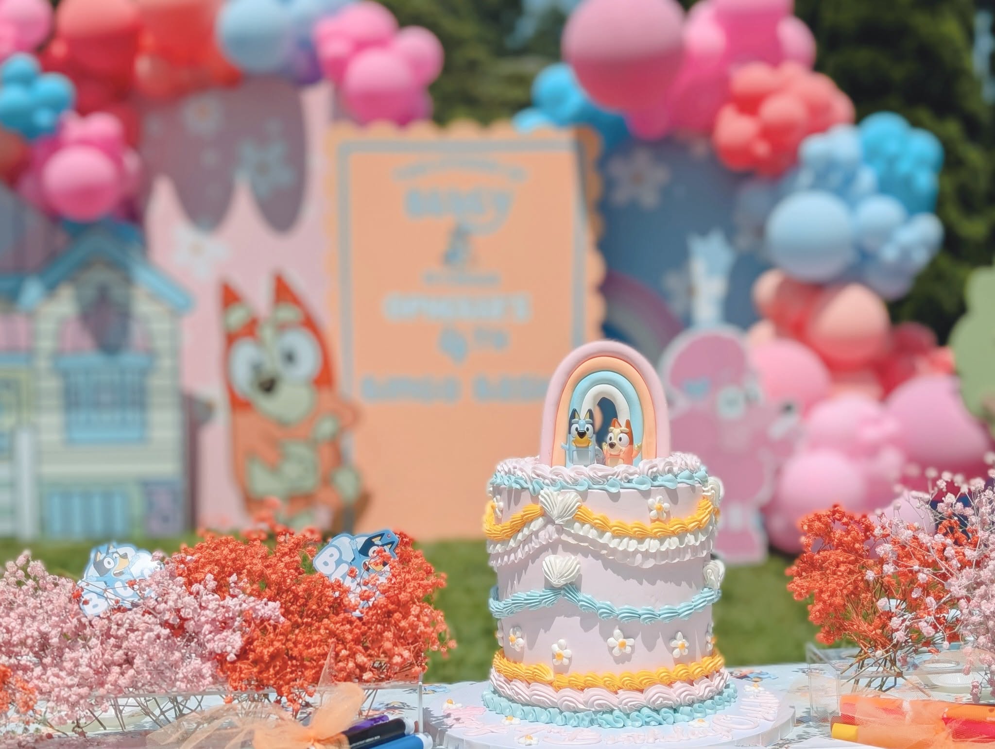 Bluey and Bingo-themed birthday cake with a rainbow topper at a party with balloon arches.