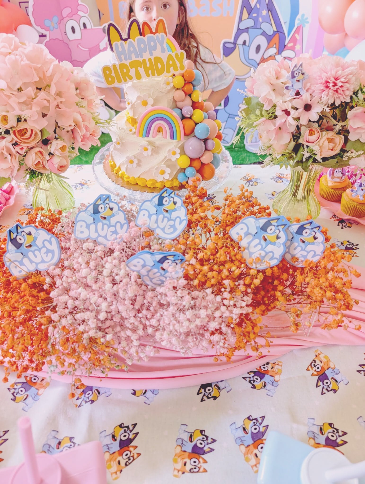 Bluey-themed birthday cake with rainbow and flower decorations, surrounded by floral centerpieces and character decor.