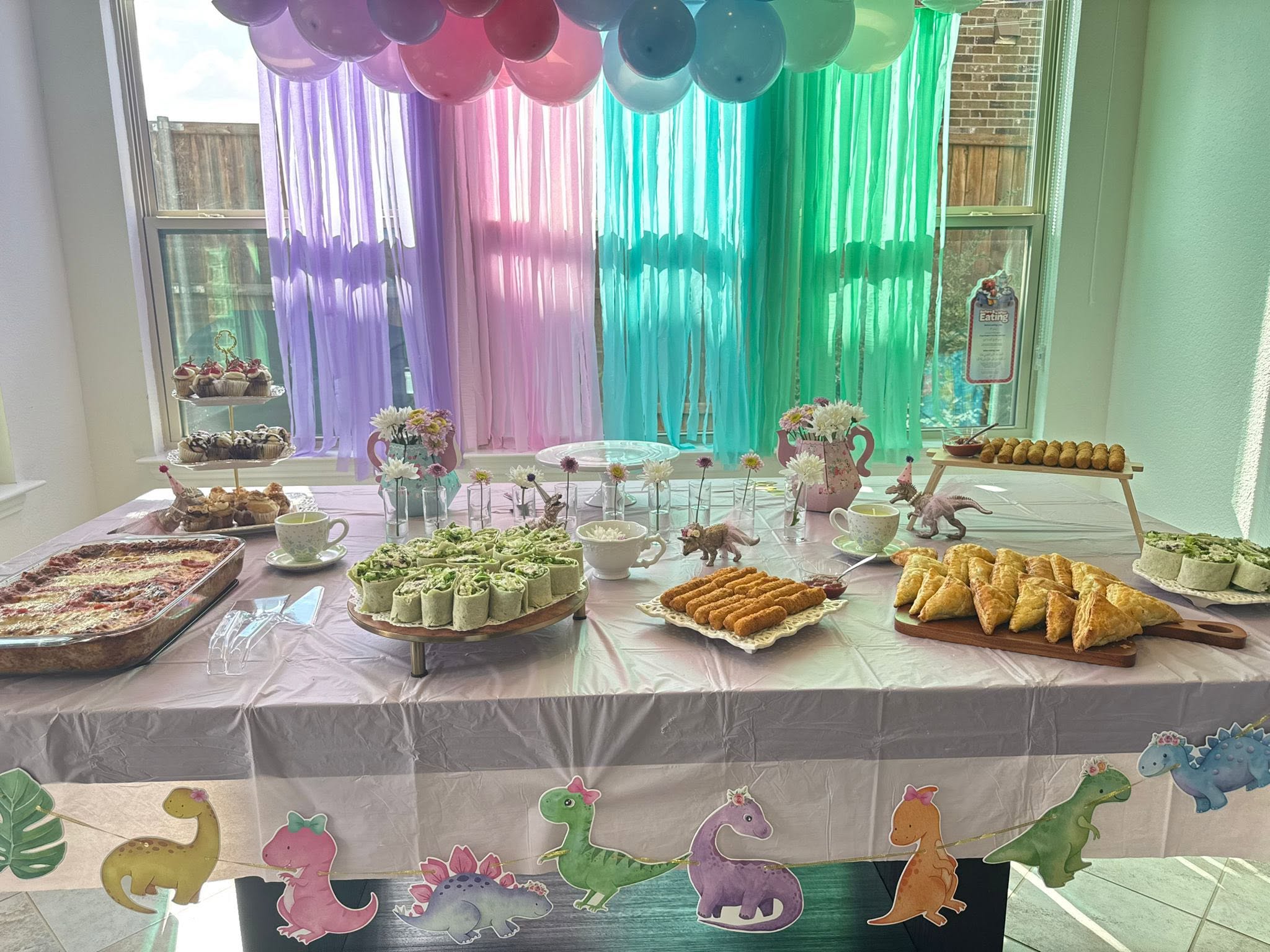 Dinosaur-themed tea party table spread with assorted finger foods, cupcakes, and pastel streamers.