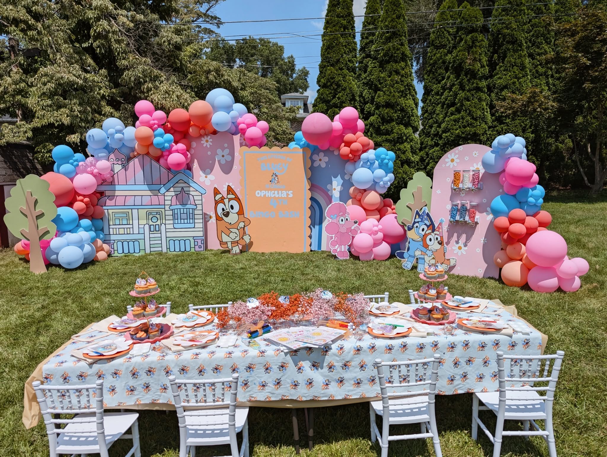 Bluey-themed birthday party setup with balloon arches, character cutouts, and a decorated table on a lawn.