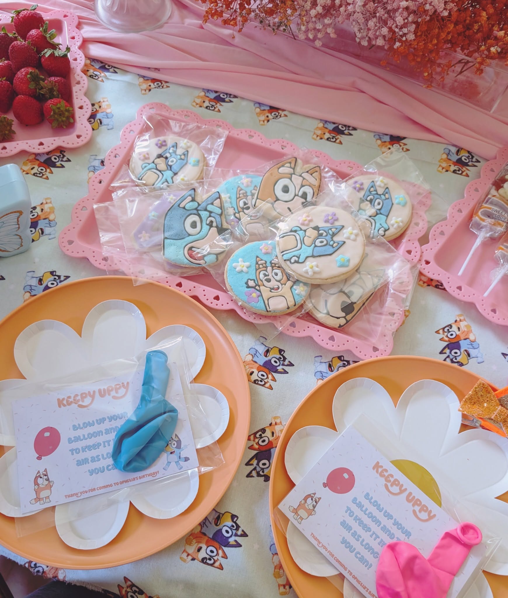 Bluey-themed party favors including decorated sugar cookies and Keepy Uppy balloon games on floral-shaped paper plates.