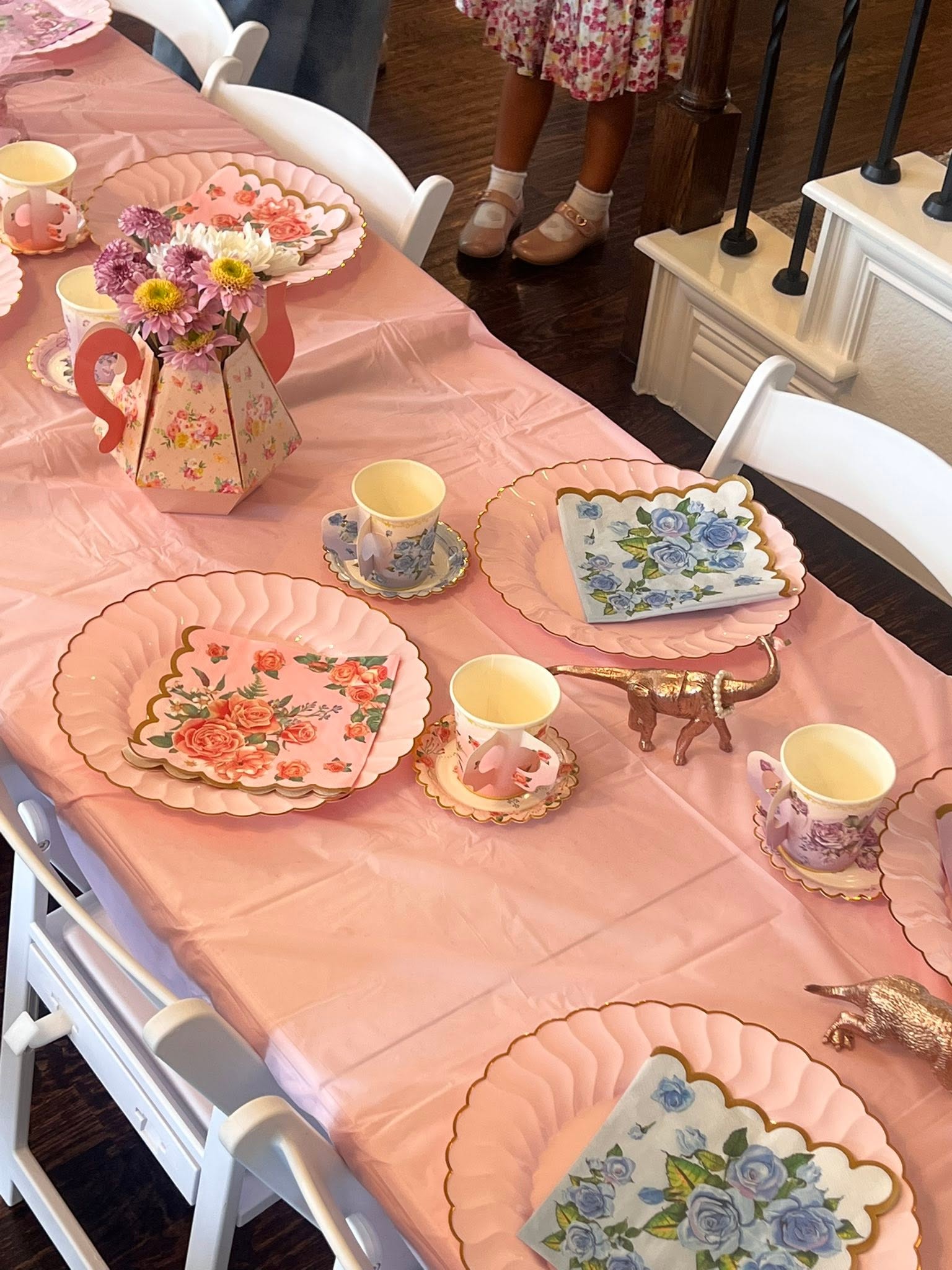 Table set for a tea party with pink tablecloth, floral paper plates, tea cups, and gold dinosaur figurines.