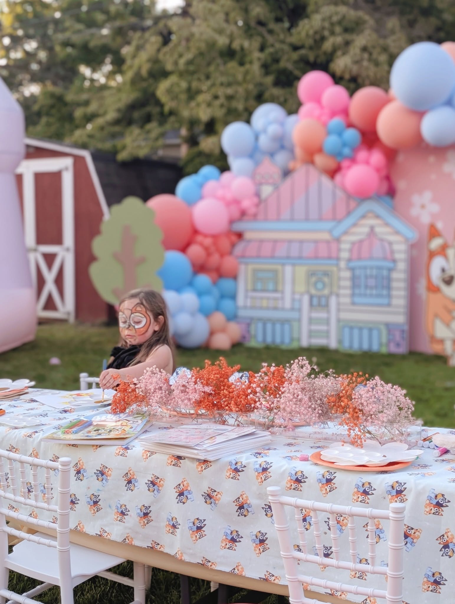 Child with face paint sitting at a Bluey-themed party table outdoors with colorful balloon decorations.