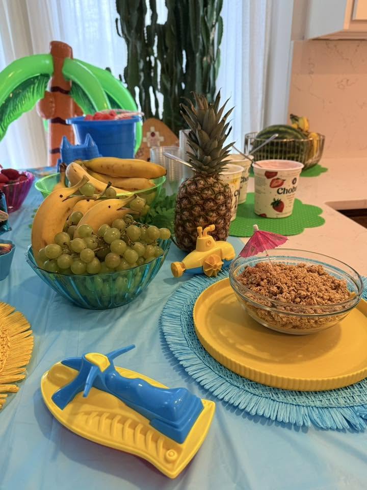 Table set for a summer themed brunch with fresh fruit bowls, granola, and tropical decorations.