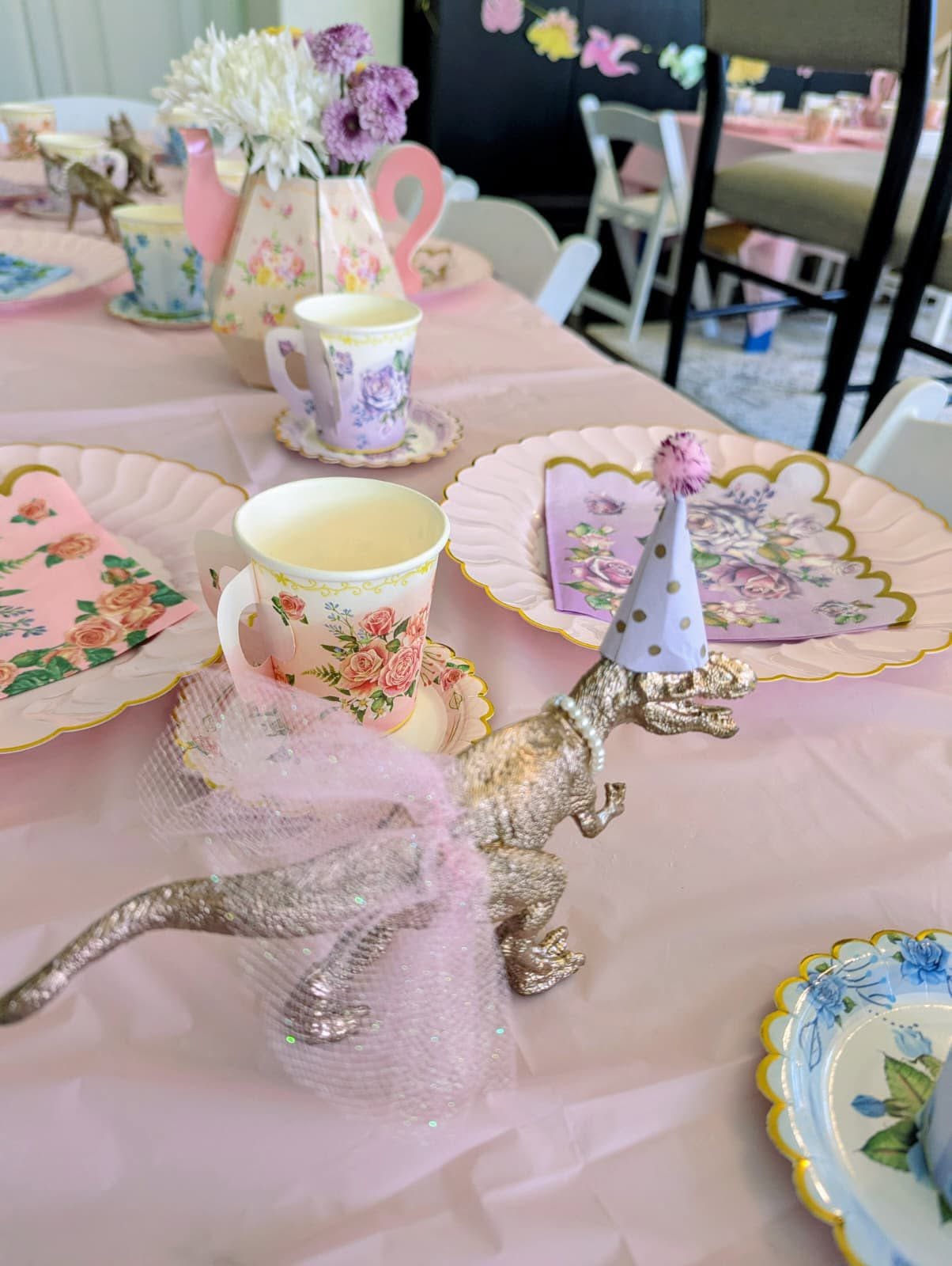 Table setting with floral teacups and a gold dinosaur figurine wearing a tutu and party hat for a party.