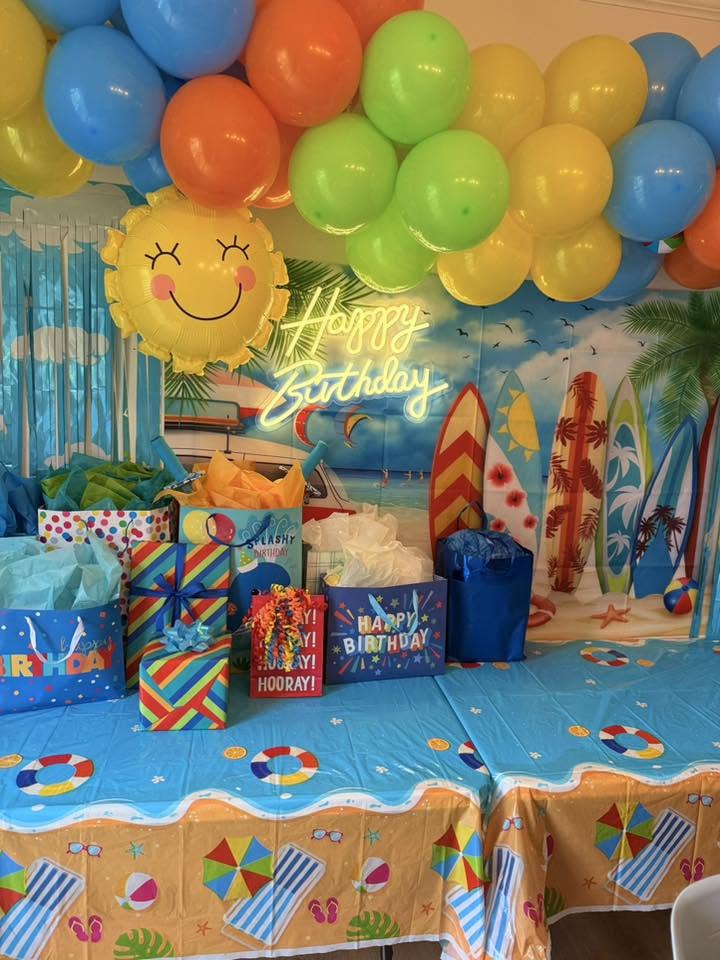 Table arranged with birthday gifts, topped with colorful balloons and a neon Happy Birthday sign against a beach backdrop.