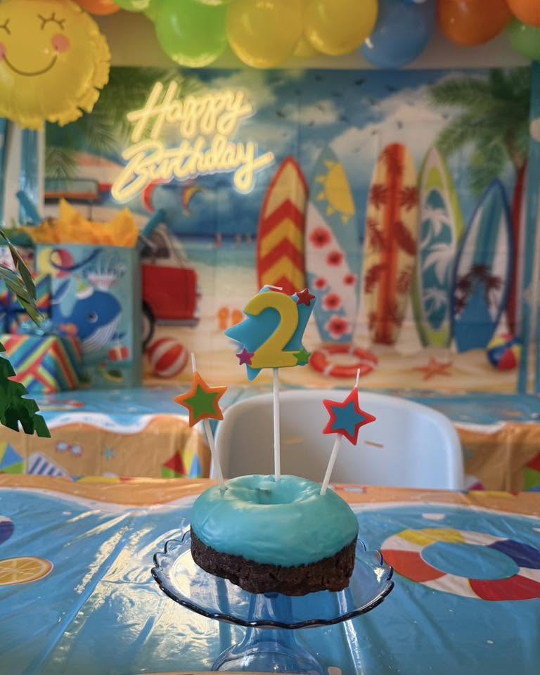 Blue glazed donut topped with a number 2 candle and star decorations, centered on a beach-themed birthday table.