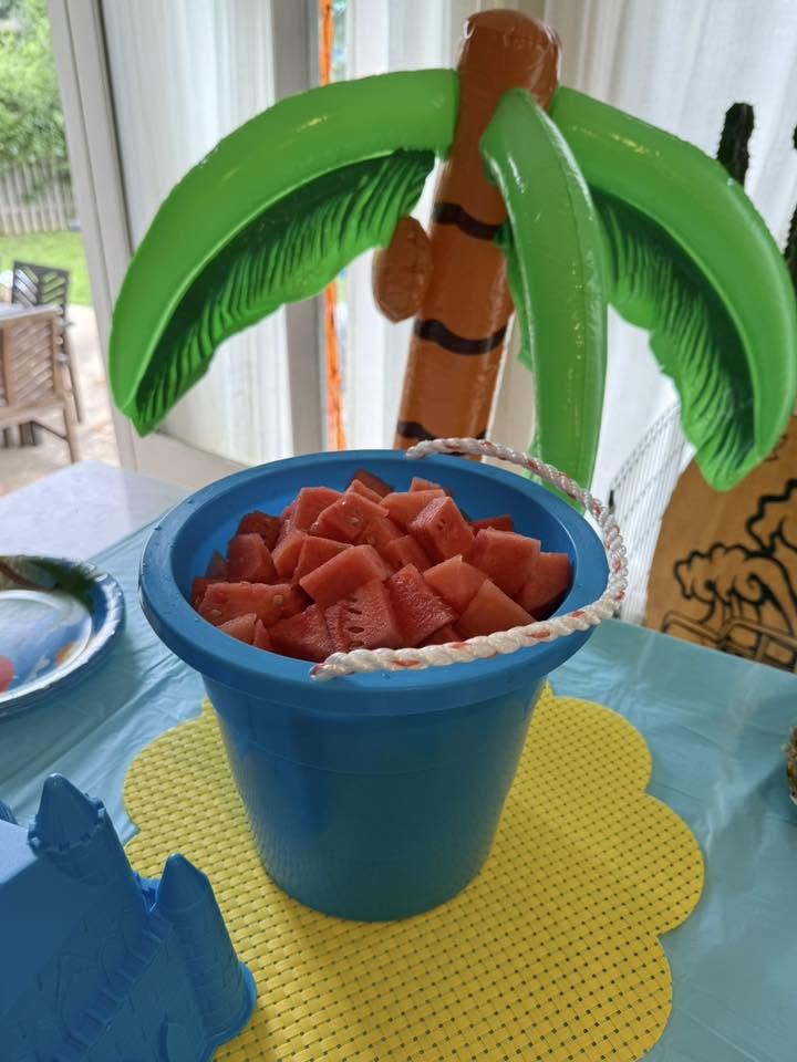 Blue plastic bucket filled with cubed watermelon sitting on a yellow placemat with an inflatable palm tree nearby
