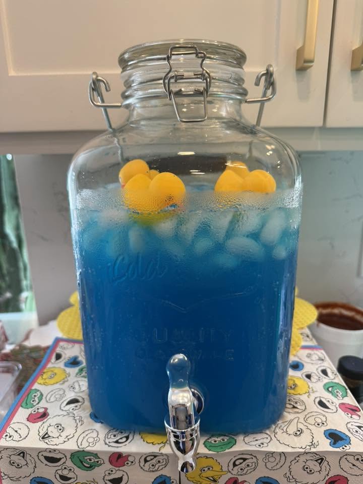 Glass beverage dispenser filled with blue punch, ice, and floating yellow rubber ducks.