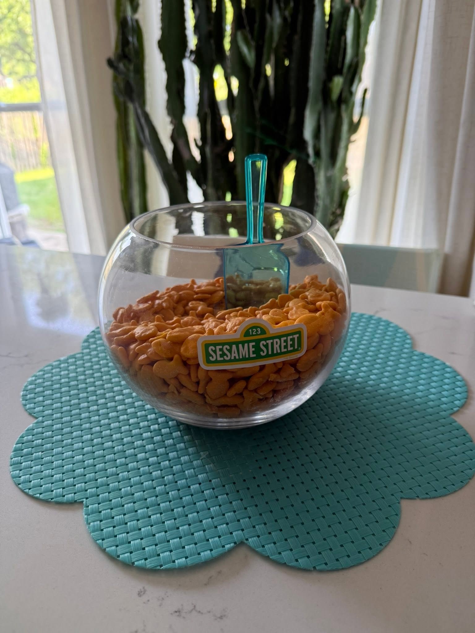 Glass bowl filled with goldfish crackers featuring a Sesame Street label and a small blue plastic scoop.