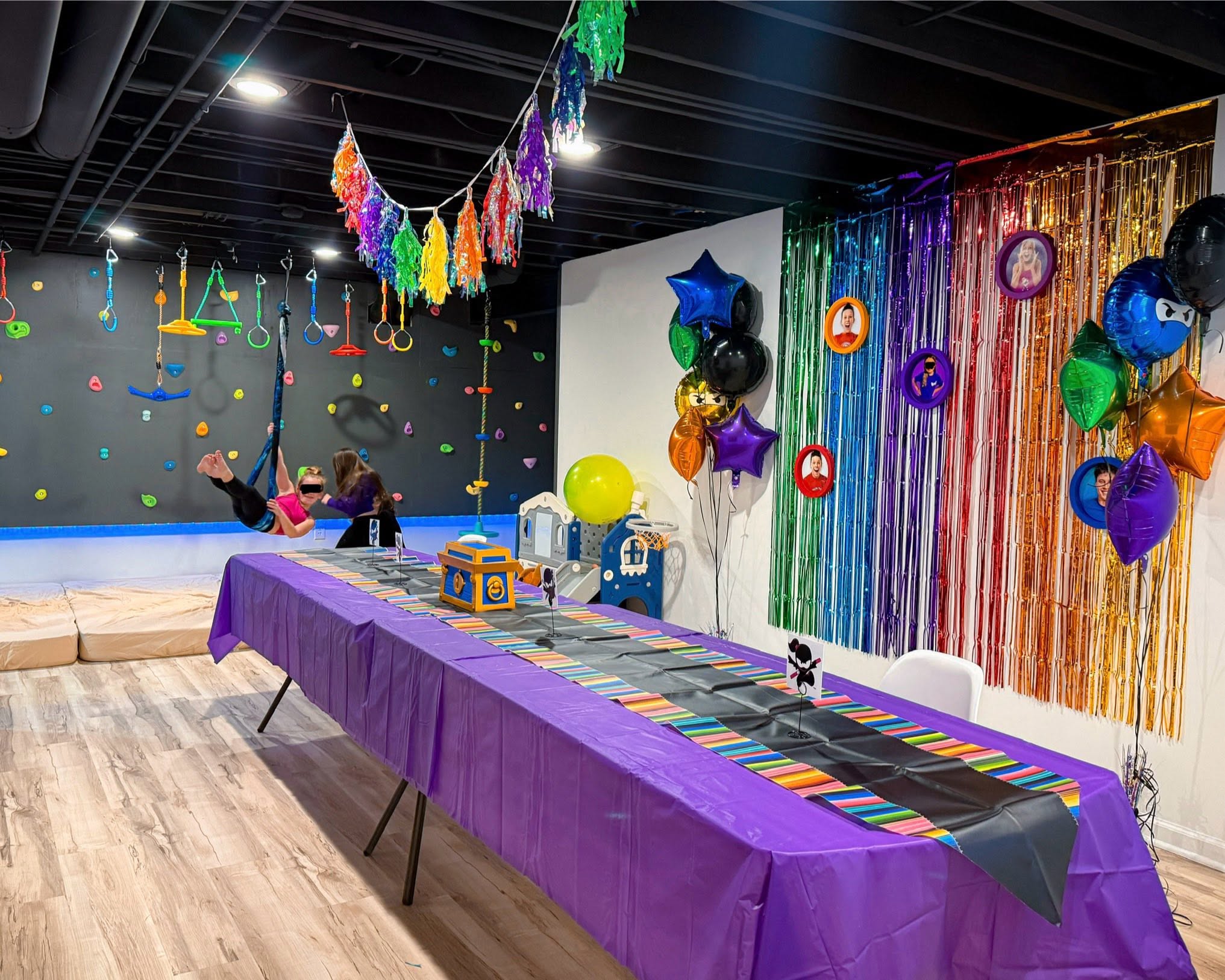 Kids playing in an indoor party space with a long dining table, climbing wall, and rainbow party decorations.