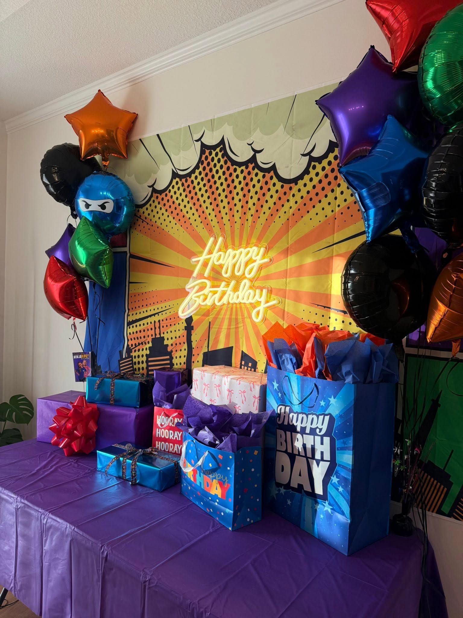 Comic book style birthday party setup with themed backdrop, neon Happy Birthday sign, and colorful balloon clusters.